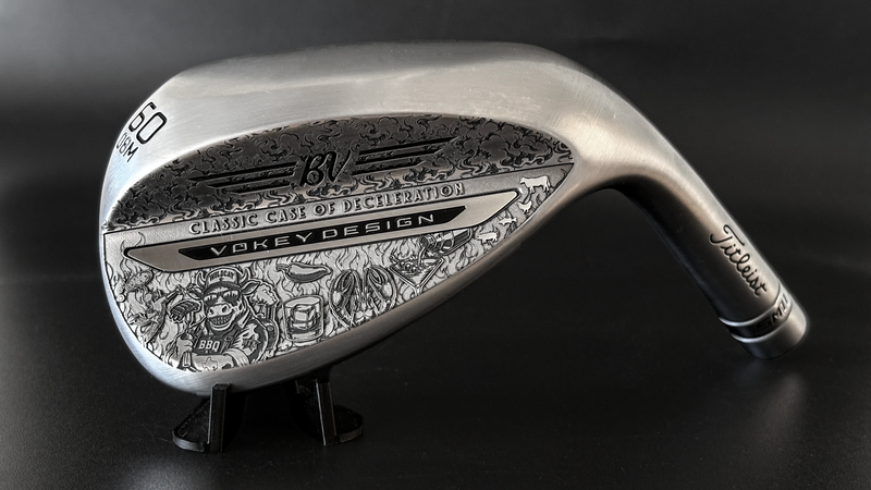 A silver Titleist Vokey golf club with intricate black engraved illustrations, displayed on a small black stand against a dark background.